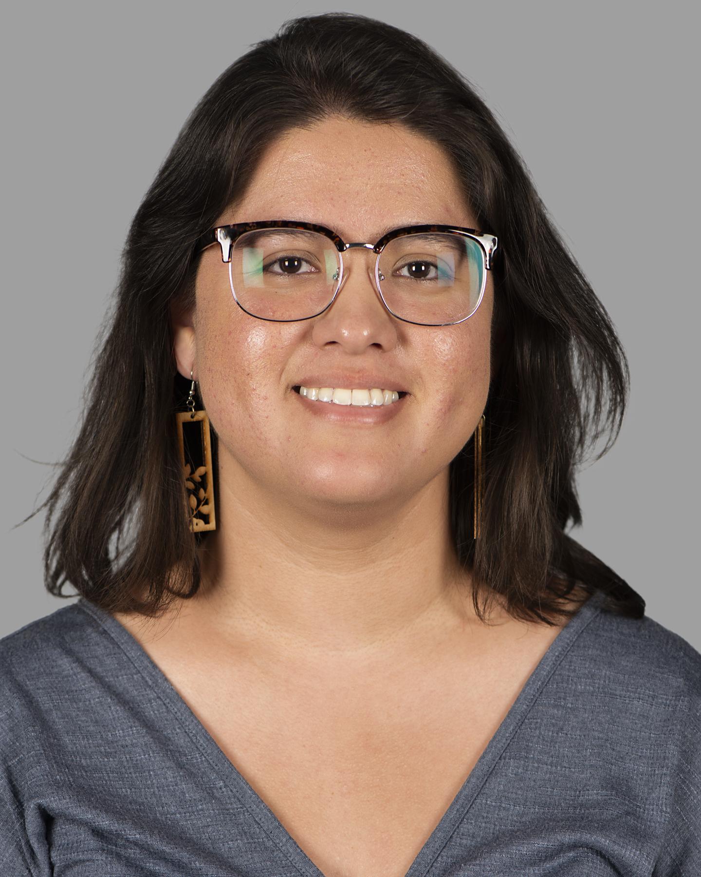 An adult with light brown skin and shoulder-length black hair wears square framed glasses and rectangular dangling wood earrings; they smile at the camera
