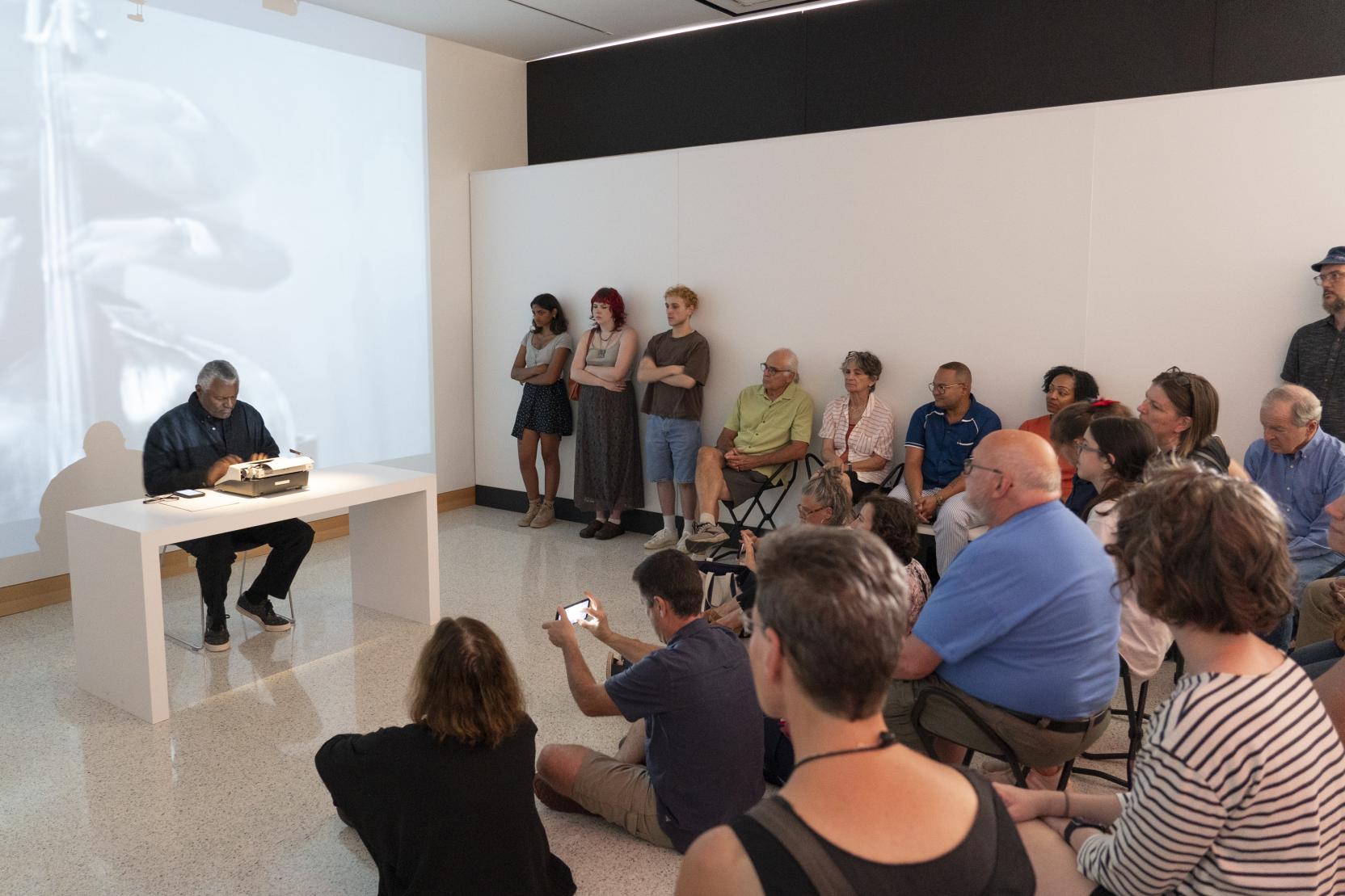 An older Black man sits at a typewriter while a video plays behind him; an audience sits in front of him