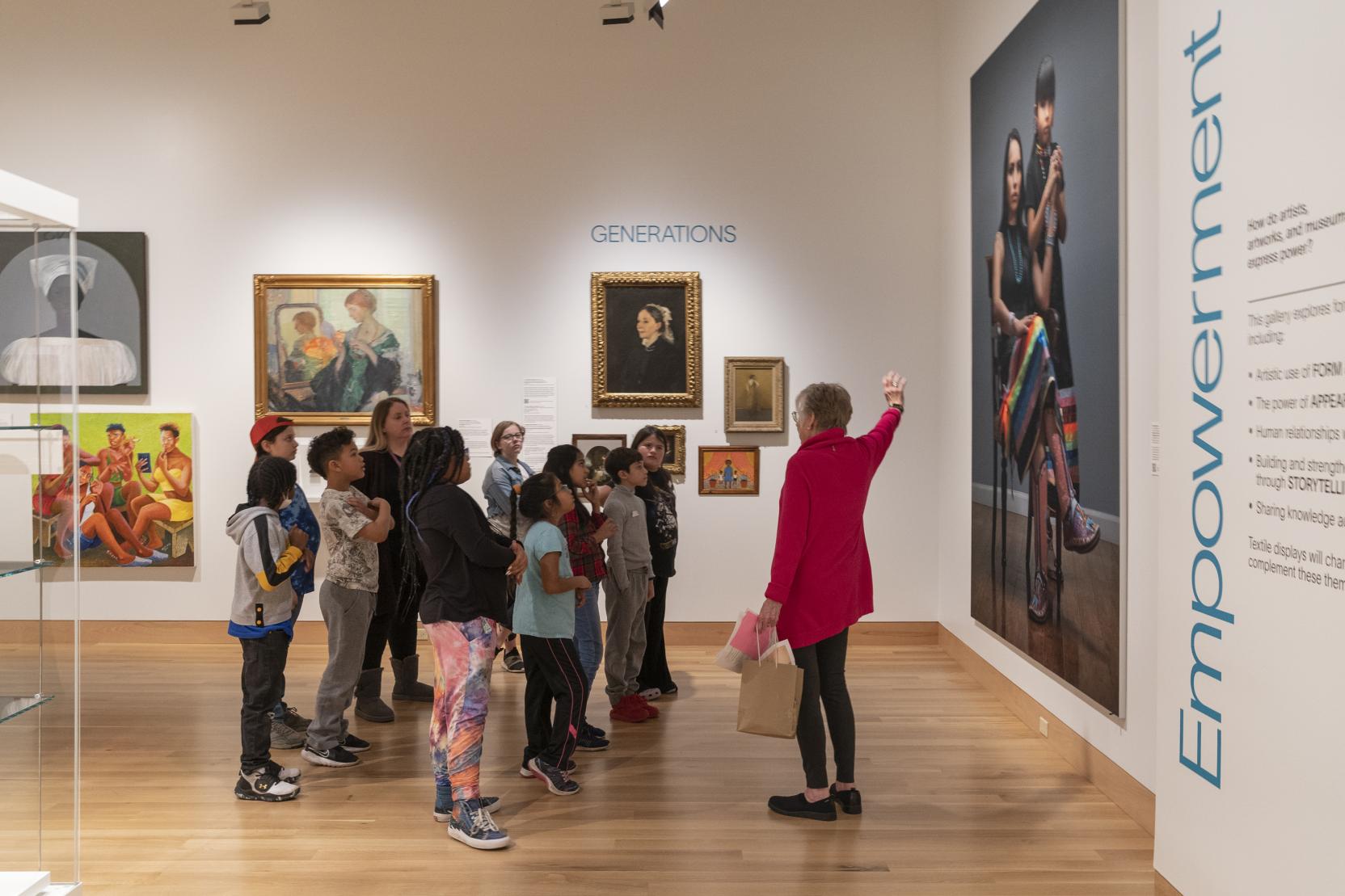 An older woman wearing a red sweater gestures toward a large portrait while a group of schoolchildren look at her