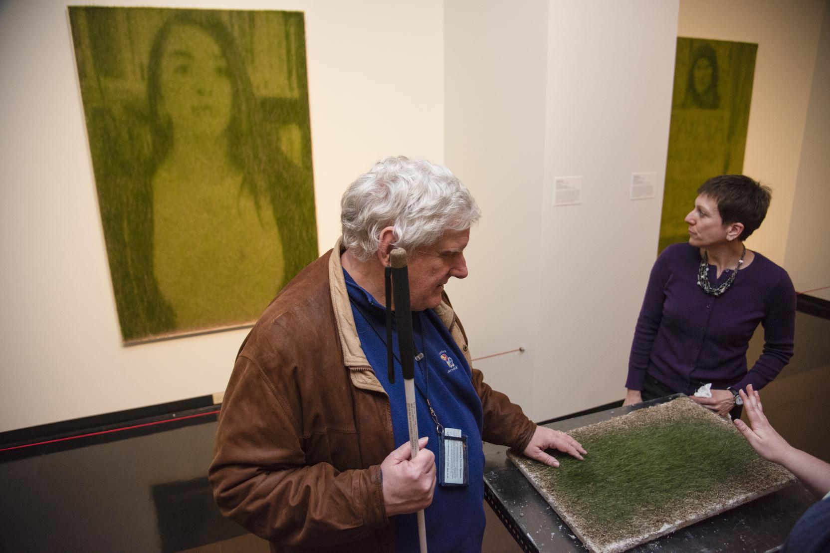 An elderly man holding a cane touches a painting on a table, a woman stands on the other side of the table