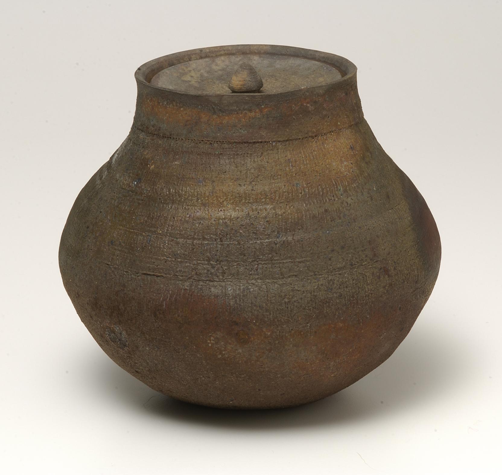 Unglazed lidded jar in brown, red, and grey hues, with a sloping shoulder, bulging body, and tapered base