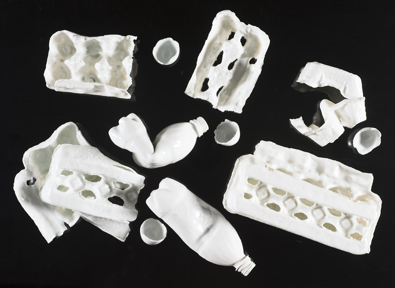 Scattered white ceramic forms of broken and intact egg cartons, squeezed plastic bottles, and broken eggshells